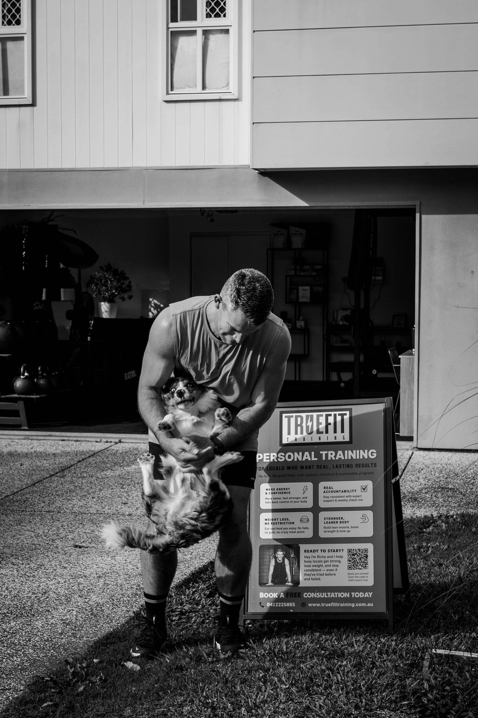 Black and white photo of Richy with his Border Collie beside the True Fit Personal Training sign outside the gym