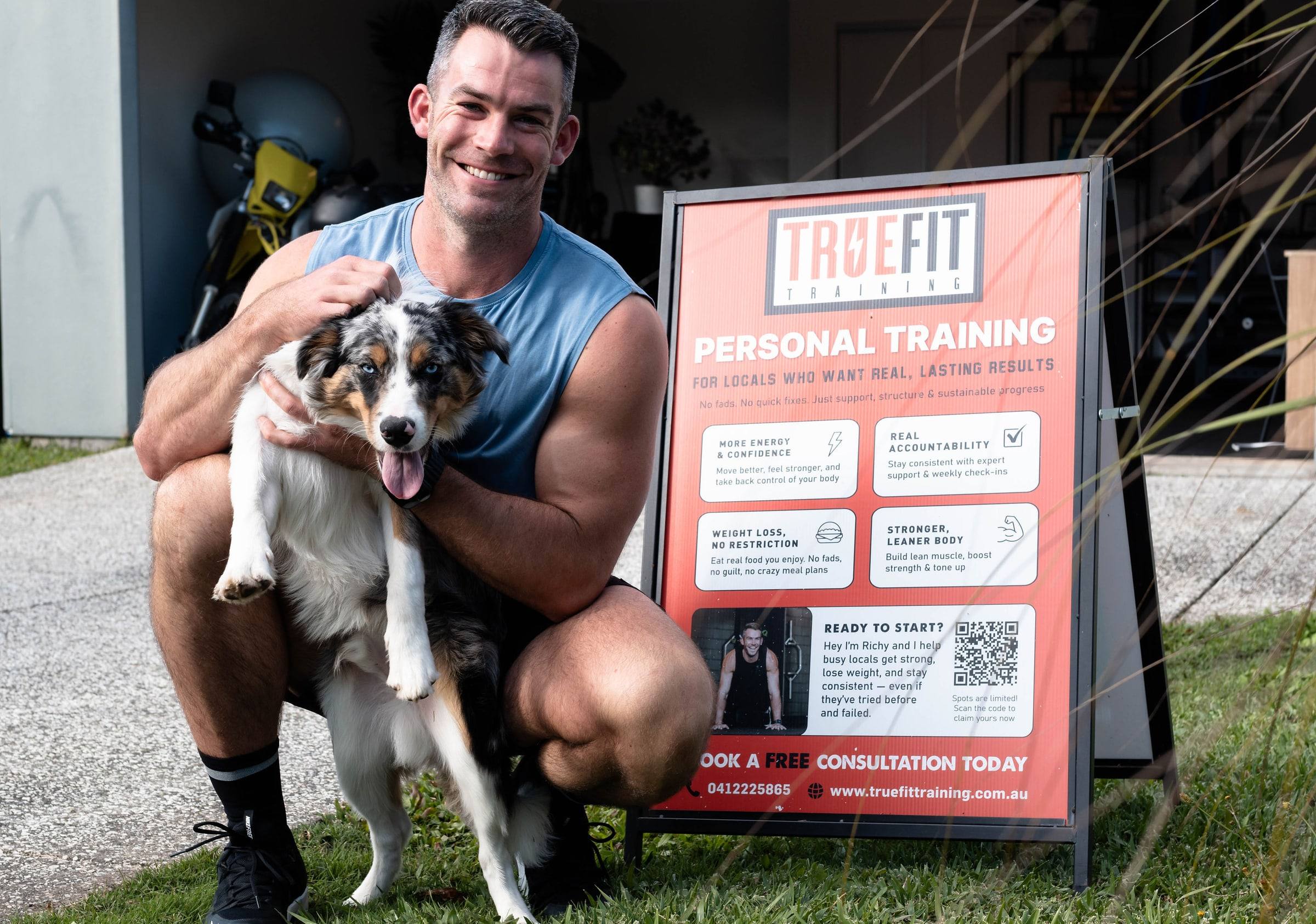 Richy crouching outside the gym holding his Border Collie, next to the True Fit Personal Training sign